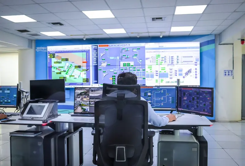 Operator at a control-room desk in front of a wall of SCADA screens